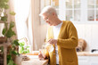 © Prostock-studio - Attractive grandmother in casual outfit pouring spring water into glass in kitchen, copy space. Side view of grey-haired senior woman drinking water while cooking, home interior