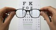 © FAWF STUDIO - Glasses held by two hands in front of a blurry Snellen chart, sharp and clear vision through the lenses showing black letters and numbers, white background, vision and clarity concept