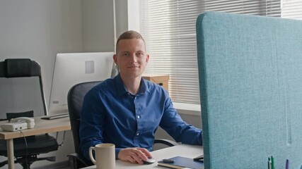 Wall Mural - Portrait of smiling adult male office worker dressed in blue shirt turning to camera while using computer at working desk in contemporary workspace, slow motion