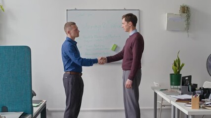 Wall Mural - Side view of two male colleagues giving handshake confirming weekly work plan outlined on whiteboard while successfully collaborating during meeting at office space