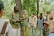 © AnnaStills - Middle aged Black man teacher holding laptop engaging with diverse group of children and teenagers standing in park listening attentively during outdoor educational activity
