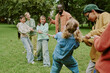 © AnnaStills - Group of multiethnic children playing tug of war outdoors with Black male teacher supervising, kids smiling and laughing while pulling rope together in green park setting