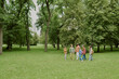 © AnnaStills - Group of multiethnic children and young Caucasian woman teacher walking together in green park, teacher leading children through grassy area surrounded by tall trees during outdoor activity