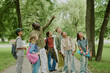© AnnaStills - Group of multiethnic children standing outdoors with Black male teacher pointing at tree, kids looking up and listening attentively, backpacks and skateboard visible, park setting with greenery