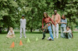 © AnnaStills - Group of diverse children playing soccer with multiethnic young adult male teacher supervising in park, kids kicking ball between cones while others watching and waiting for turn on grass
