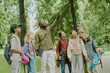 © AnnaStills - Black middle aged man teacher leading group of diverse children outdoors in park, teacher pointing at tree while multiethnic kids with backpacks and skateboard looking up and listening attentively