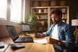© Stockphotodirectors - A man sits at a wooden desk in a well-lit home office, engaged in writing notes while drinking from a cup. He is surrounded by a laptop, a tablet, and house plants, indicating a productive workspace.