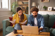 © Stockphotodirectors - A couple sits together on a couch in their living room, with one partner reviewing life insurance documents while the other works on a laptop. The atmosphere is relaxed and productive.