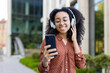 © Liubomir - A young Black woman wearing glasses and headphones smiles while looking at her smartphone outdoors near a modern building.