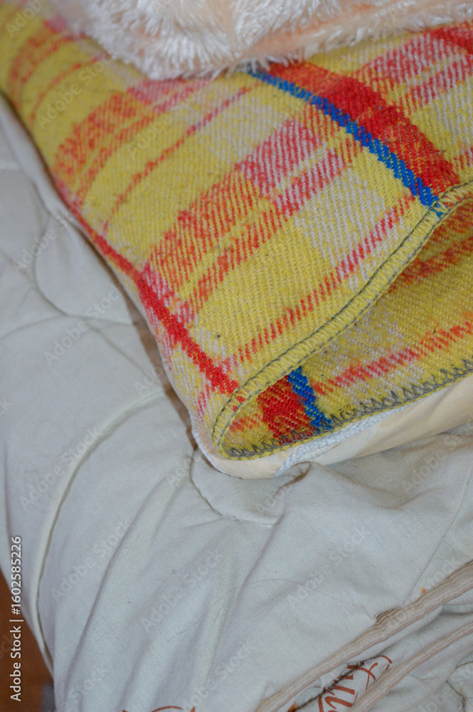 a close-up of a cozy bed scene with layers of different blankets and textiles. a soft peach faux fur blanket is placed over a yellow and red checkered woven throw, all resting on a light-colored fabri
