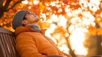 Wall Mural - Young man relaxed enjoying autumn leaves on a park bench with warm sunlight background