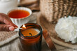 © shine.graphics - Hand lighting a candle in a cozy flat lay scene with tea, white peonies, straw hat, and an open book. Warm summer moment of relaxation and self-care in soft natural light