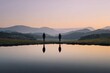 © Both - Two figures stand facing a tranquil mountain vista mirrored in a serene lake at dawn