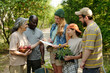 © pressmaster - Group of multiethnic adults and teenager gathering outdoors, smiling and interacting while holding apples, clipboard, and potted flowers, standing in green park setting