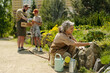 © pressmaster - Senior Caucasian woman tending garden bed while middle aged Caucasian man and teenage boy gathering vegetables in background, outdoor community garden setting, gardening tools visible