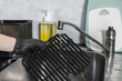 © Alina - Hand wearing a black protective glove washes a dirty non-stick air fryer grill grate under running kitchen water, illustrating appliance care and home hygiene