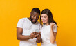 © Prostock-studio - Mobile bonus. Surprised multiracial couple looking at smartphone screen and exclaiming with joy, posing together on yellow background, free space