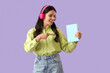 © Pixel-Shot - Beautiful woman in headphones pointing at book on lilac background