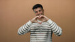 © Krakenimages.com - Young man smiling while making a heart shape with hands against a brown background, wearing a striped shirt, conveying happiness and warmth in a simple portrait.