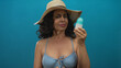 © Krakenimages.com - Woman wearing a hat and bikini examines lotion bottle against isolated blue background, exuding an inquisitive expression while maintaining a relaxed summertime vibe.