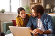 © Stockphotodirectors - A couple is engaged in a serious conversation about their financial challenges while sitting together on a couch, focused on a laptop. The atmosphere suggests concern.