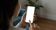 © Dual Studio - Young woman user holding smart phone in hand touching finger mock up white empty cellphone screen using mobile applications at home. Display mockup close up over shoulder view.