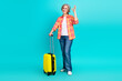 © deagreez - Smiling elderly woman with grey hair in casual clothing holding a yellow suitcase and showing an OK gesture on a teal background
