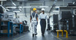 © Gorodenkoff - Diverse Female and Male Technicians Walking Through a Modern Factory, Discussing Work. Young Man Wearing a Yellow Hard Hat and Overalls, Manager Dressed in Business Attire. Footage From the Back