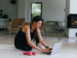 © .shock - A young woman in sportswear is sitting in the living room and preparing for online training while using a laptop.