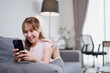 © Tj - Happy young woman is lying on the sofa and using mobile phone, browsing internet, chatting in social networks, watching videos, playing mobile games, shopping online at home