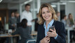 © Pete - Mature businesswoman smiling confidently holds smartphone in modern office setting. Colleagues blurred in background, indicating professional workplace. Focus on leader, conveying success,