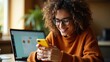© MT Studio - Woman with curly hair and glasses smiling while using a yellow smartphone next to a laptop computer