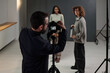 © TrueFrame Collective - Caucasian man operating camera on tripod photographing Black young adult woman and Caucasian woman standing in studio during acting audition