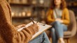 © Jiraphat - A person wearing a brown sweater takes notes on a clipboard during a therapy session with a woman sitting on a chair in the background.
