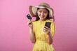© Daniel - Asian woman wearing a summer hat and yellow dress smiling while holding a credit card and smartphone, standing in front of a pink background