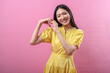 © Daniel - Asian woman in a yellow dress forms a heart shape with her hands while smiling warmly. She stands against a bright pink background, radiating positivity, affection, and cheerful energy