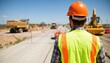 © Raisa - Construction worker in orange hard hat, vest stands on site holding clipboard with detailed drawing of ongoing road construction. Large yellow vehicle, orange excavator visible in background with