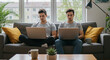 © AswinRizky - Remote Work Double, Young Men On Couch Working Remotely on Laptops In Bright Living Room , Twin Brothers Work From Home