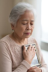 Wall Mural - close-up of elderly east asian woman holding photo frame close to heart soft home light and gentle s