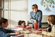 © DragonImages - Caucasian young adult man teaching diverse group of children about plant biology using strawberry model, children listening and taking notes at classroom table during science lesson