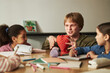 © DragonImages - Caucasian young adult man with strawberry model explaining plant growing to diverse group of children sitting around table, children listening attentively and engaging in educational discussion