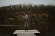 © My Ocean studio - Woman in a gray coat and beanie stands on a wooden pier, holding a wine glass, facing a calm river and rocky forest shoreline under an overcast sky.