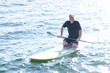 © Mat Hayward - Happy middle aged man resting on his knees on a paddle board outside on a lake. He is holding a paddle over the water and working to maintain balance.