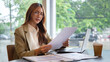 © wattana - Confident young businesswoman reading documents while working on a laptop at a modern cafe.