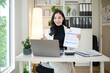 © wattana - Cheerful businesswoman conducting an online meeting, holding a report with graphs and financial data in a modern home office.