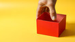 © Cynthia - Mano sosteniendo una caja roja de carton roja sobre un  fondo amarillo liso y aislado. Vista de frente. Copy space