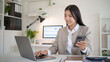 © matinee - Smiling businesswoman using her phone while checking data on a laptop at office desk.