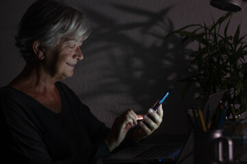 Modern elderly woman using phone while working on computer at night. Attractive businesswoman studying online, using laptop software, web surfing information or shopping in internet store