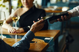 Caucasian young adult woman sitting at restaurant table, holding wine glass while Caucasian young adult man making contactless payment with smartphone to waiter holding terminal