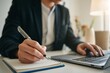© Ajay - Businesswoman writing notes in notebook while using laptop, productivity, planning, remote office workspace.